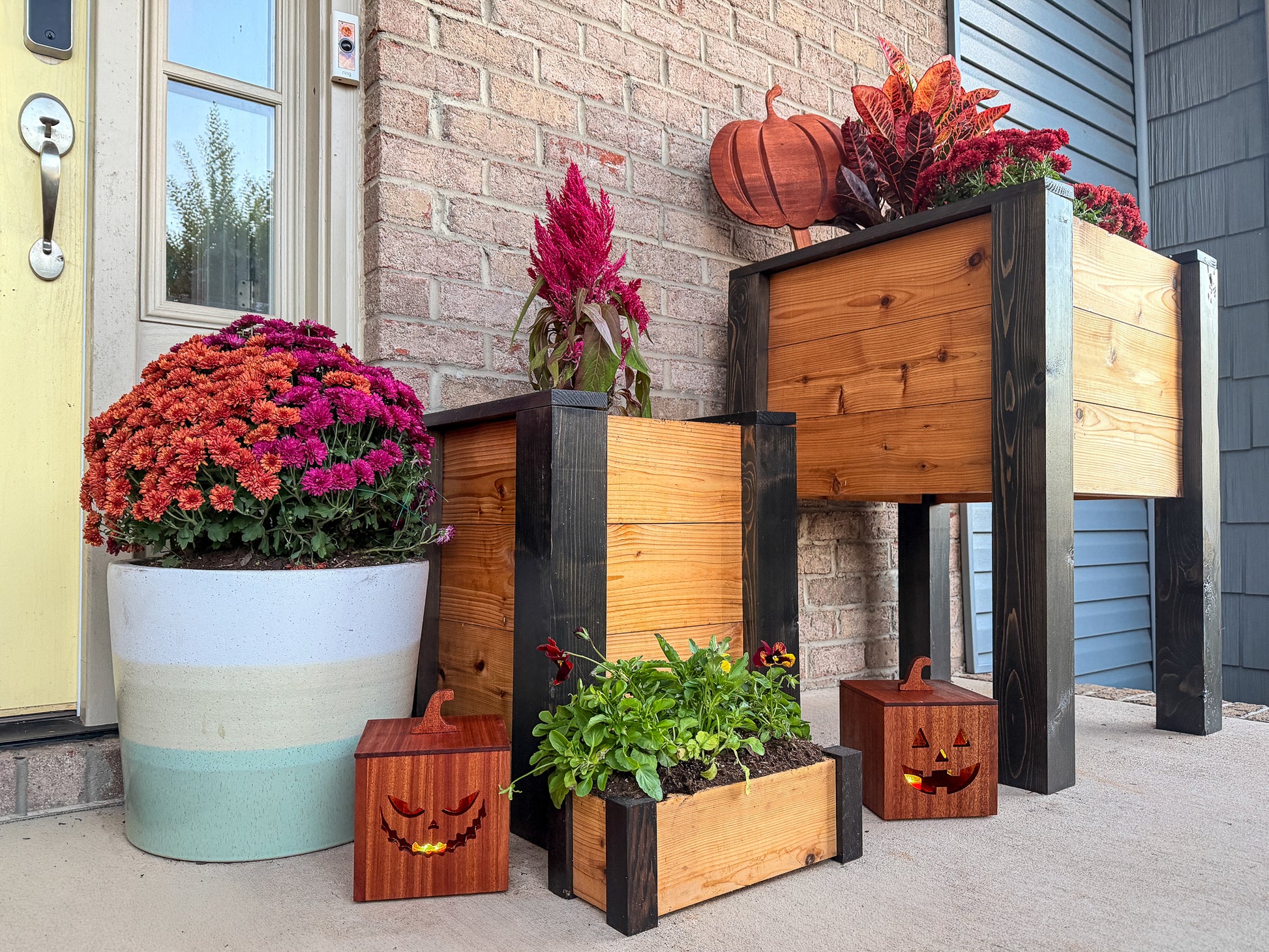 Decorative outdoor raised garden beds with wood jack-o-lanterns, pumpkins, and flowers on the front porch of a house.
